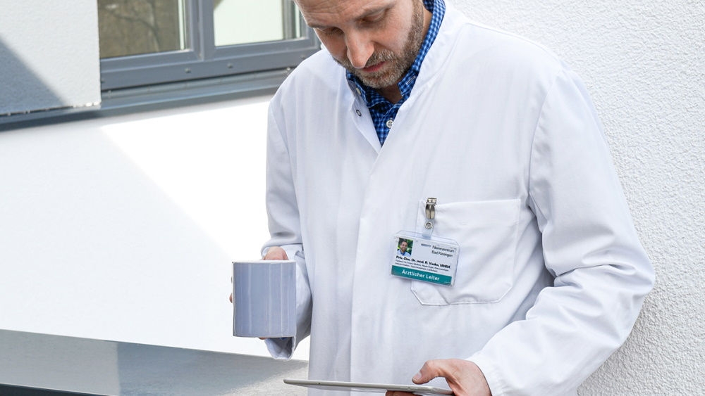 Ein männlicher Arzt hält eine Tasse Kaffee und arbeitet mit seinem Tablet