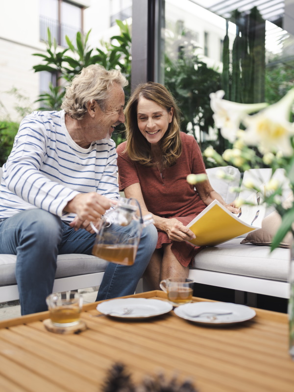 Ein Mann mit einer Teekanne und eine Frau mit einer Zeitschrift sitzen auf einem Sofa und lachen