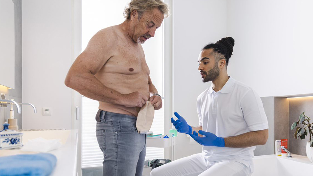 Ein Mann mit nacktem Oberkörper im privaten Badezimmer manipuliert einen Stomabeutel auf seinem Bauch. Eine männliche Pflegekraft sitzt auf dem Badewannenrand und gibt Anweisungen.
