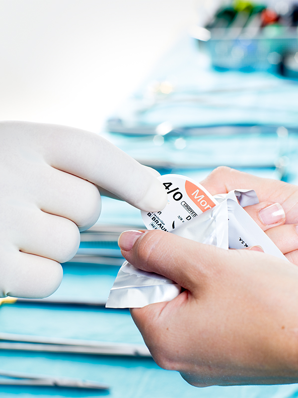 Hand mit Handschuh entnimmt Nahtmaterial aus einer sterilen Verpackung