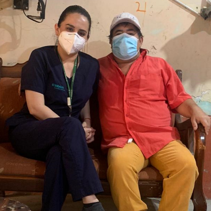 Carol sitzt neben Carlo auf einem Sofa, schaut in die Kamera und trägt eine Gesichtsmaske.